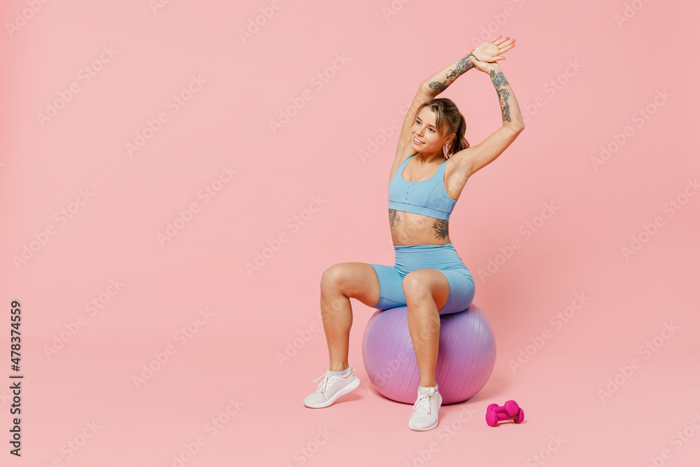 Full body young athletic fitness trainer woman in blue tracksuit spend time in home gym sit on fitball tilt body sides with hand stretch lunge isolated on plain pink background Workout sport concept Full body young athletic fitness trainer woman in blue tracksuit spend time in home gym sit on fitball tilt body sides with hand stretch lunge isolated on plain pink background Workout sport concept