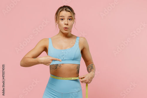 Young shocked sporty athletic fitness trainer instructor woman wear blue tracksuit spend time in home gym point finger on measure tape on waist isolated on plain pink background Workout sport concept