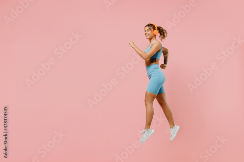 Full body young strong sporty fitness trainer instructor woman wear blue tracksuit headphones spend time in home gym jump high run isolated on pastel plain light pink background Workout sport concept