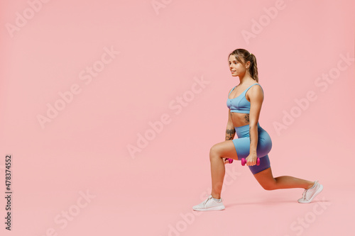 Full size young sporty athletic fitness trainer instructor woman wear blue tracksuit spend time in home gym hold dumbbells do stretch squats isolated on plain pink background. Workout sport concept
