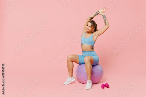 Full body young athletic fitness trainer woman in blue tracksuit spend time in home gym sit on fitball tilt body sides with hand stretch lunge isolated on plain pink background Workout sport concept