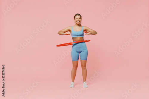 Full size young strong sporty athletic fitness trainer instructor woman wear blue tracksuit spend time in home gym using hula hoop isolated on pastel plain light pink background Workout sport concept.