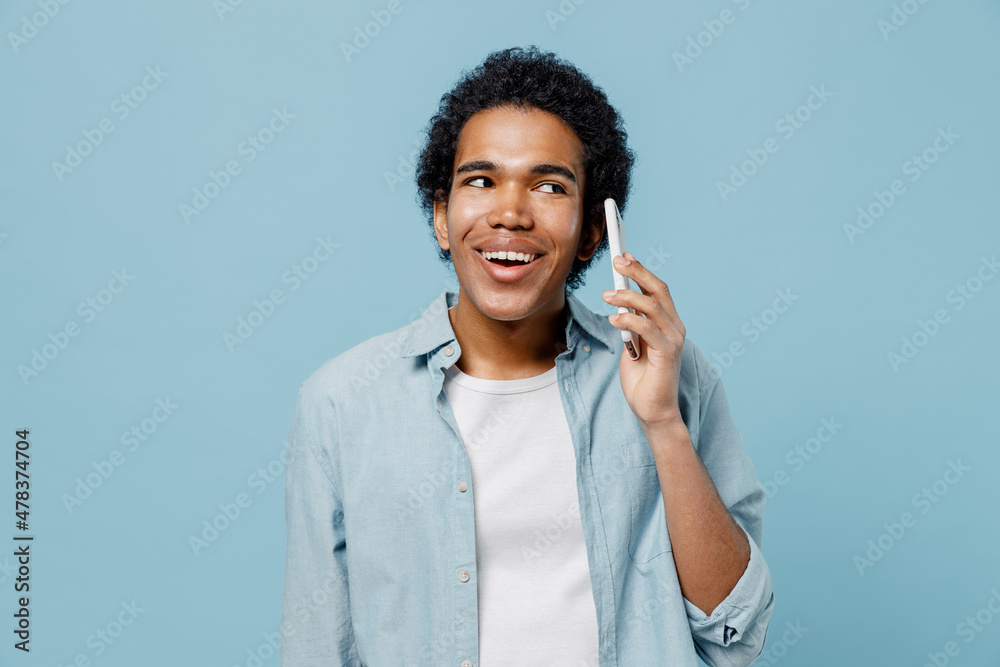 Young black curly man 20s years old wears white shirt hold in hand use talk speak on mobile cell phone conducting pleasant conversation isolated on plain pastel light blue background studio portrait. Young black curly man 20s years old wears white shirt hold in hand use talk speak on mobile cell phone conducting pleasant conversation isolated on plain pastel light blue background studio portrait.