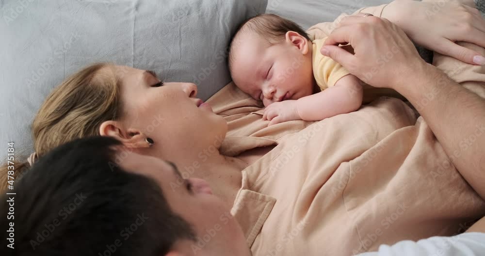 Loving parents looking at adorable baby boy sleeping in arms while lying on bed at home Loving parents looking at adorable baby boy sleeping in arms while lying on bed at home