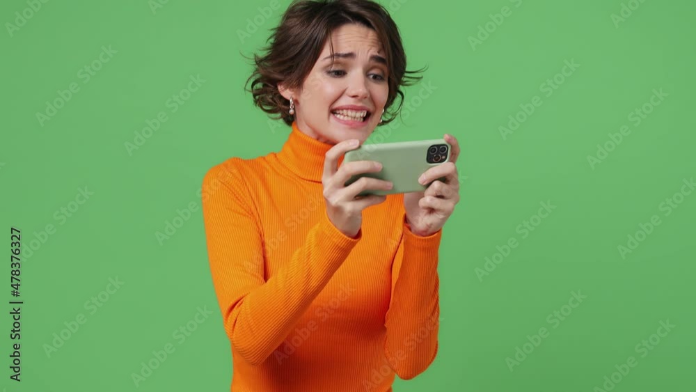 Gambling fascinating fun young brunette woman 20s wears orange shirt using play racing on mobile cell phone hold gadget smartphone for pc video games isolated on plain green background studio portrait Gambling fascinating fun young brunette woman 20s wears orange shirt using play racing on mobile cell phone hold gadget smartphone for pc video games isolated on plain green background studio portrait