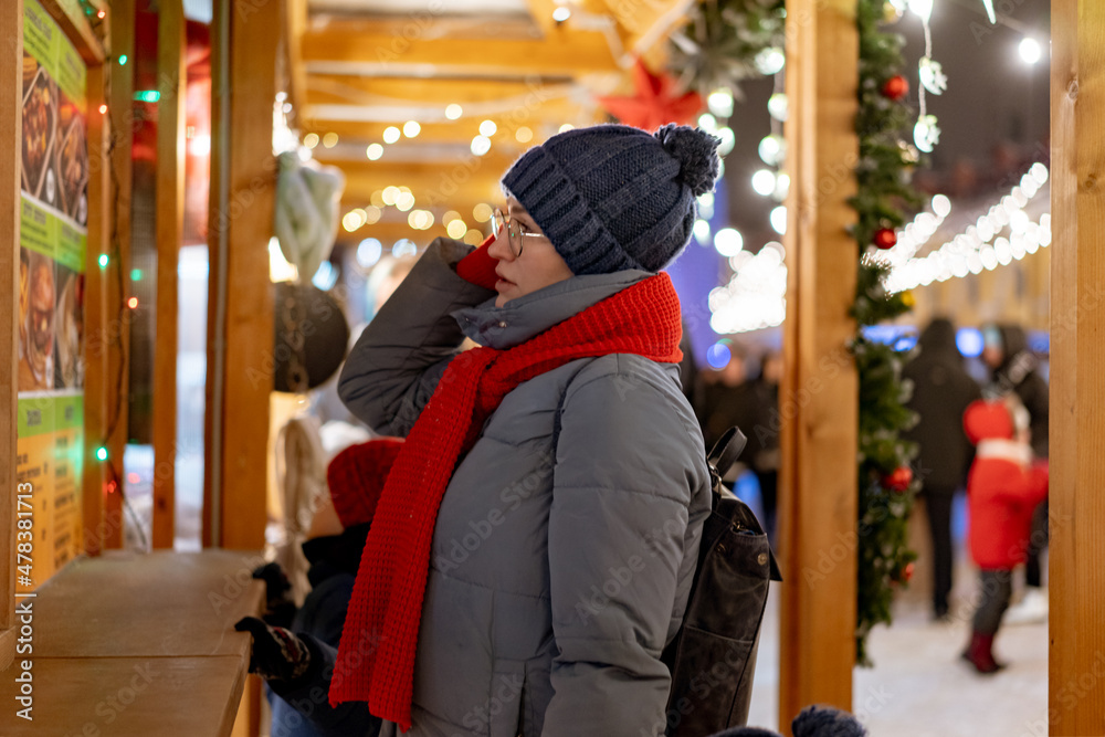 caucasian woman wearing hat and red scarf going to buy treats in kiosk at christmas market