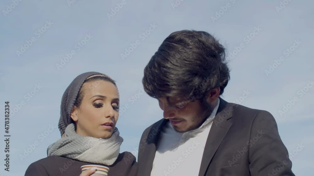 Gorgeous Arabic couple drinking coffee outdoor and talking. Happy woman wearing hijab and handsome bearded man in official suit having romantic date in city and looking at sights. Love concept Gorgeous Arabic couple drinking coffee outdoor and talking. Happy woman wearing hijab and handsome bearded man in official suit having romantic date in city and looking at sights. Love concept
