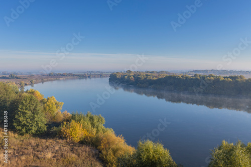 Autumn landscape in the early morning overlooking the river. A wide river and endless expanses of fields. Yellow leaves on trees and bushes are illuminated by the rays of the rising sun. © Sergei