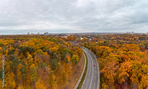 Aerial panorama, scenic asphalt road curve in autumnal forest near residential district with cloudy sky. Fly above street in autumn city park. Treetop view in Kharkiv, Ukraine