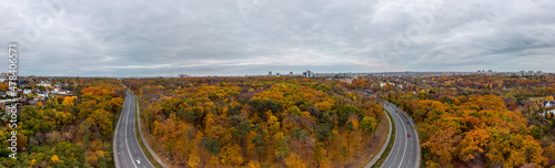 Aerial wide panorama with scenic asphalt road radial curve in autumnal forest near residential district with cloudy sky. Fly above street in autumn city park. Treetop view in Kharkiv, Ukraine