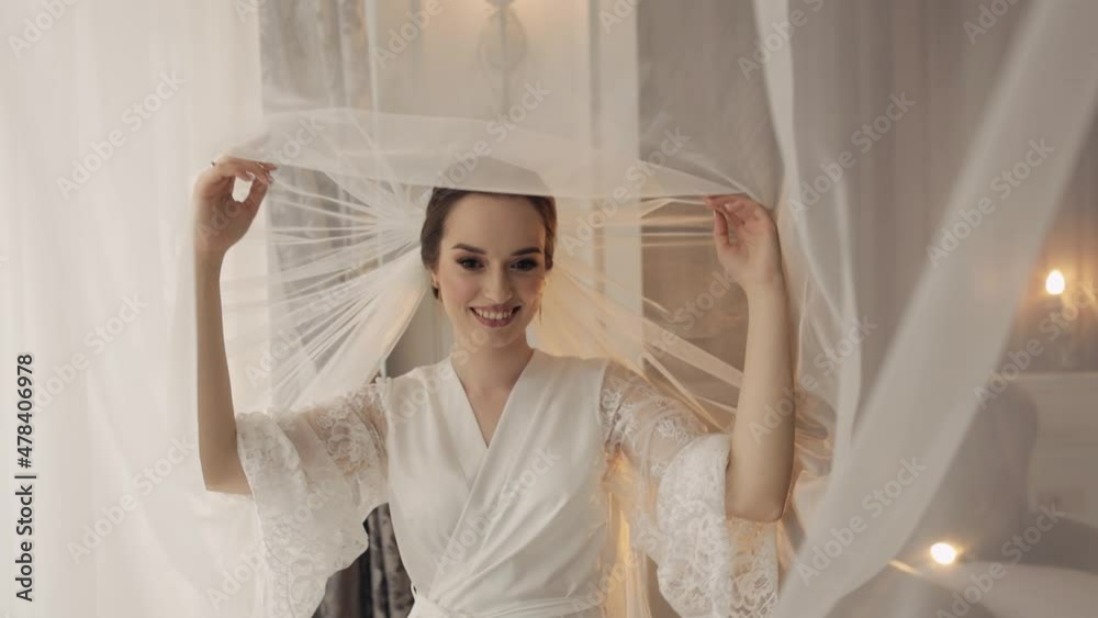 Bride in white boudoir dress under veil and in a silk robe. Wedding morning preparations before ceremony. Luxury bride with fashion hairstyle and makeup. Beautiful, lovely woman in night gown and veil Bride in white boudoir dress under veil and in a silk robe. Wedding morning preparations before ceremony. Luxury bride with fashion hairstyle and makeup. Beautiful, lovely woman in night gown and veil