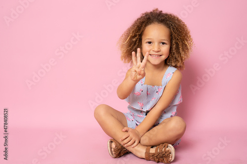 Beautiful little girl sitting on the floor counting three with fingers over pink background.