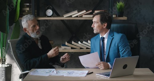 Two mature businessmen discussing business growth, looking at paper documents and using laptop computer in office, sitting at desk. Man ceo talking with colleague about deal while meeting indoors