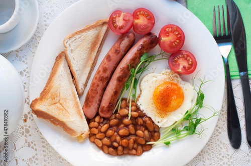 English breakfast with scrambled egg, sausages, toast, tomatoes, pea sprouts and beans on white plate. Traditional english breakfast with tea and teapot on white lace tablecloth. Top view