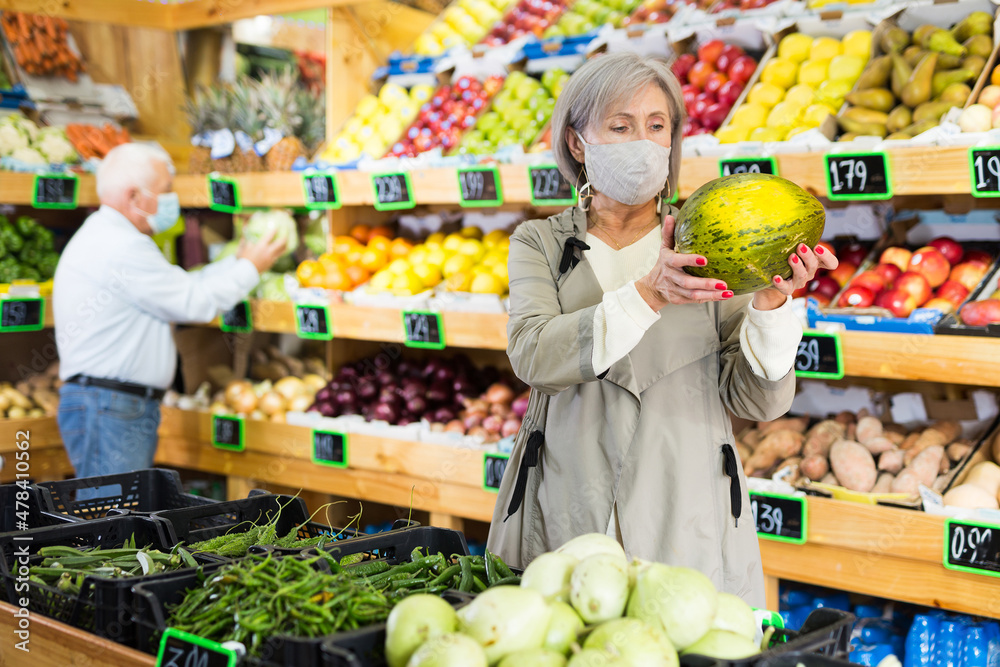 Woman in protective mask selecting watermelon in greengrocer. Man walking in background