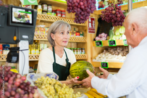 Woman supermarket employee hands watermelon to an elderly man after paying for purchase at the checkout