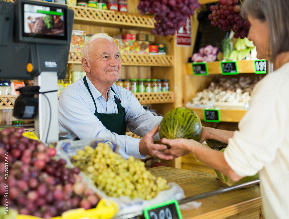 Senior salesman standing at counter in greengrocer and serving woman customer who purchasing fresh melon.