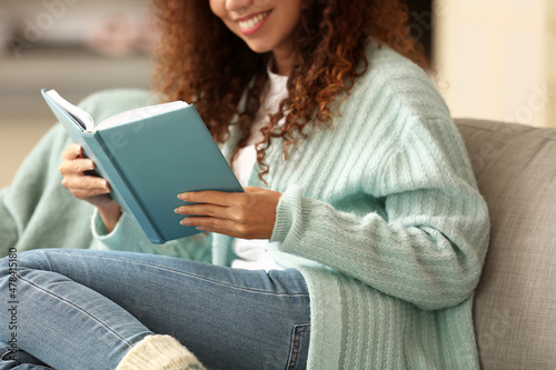 Young African-American woman in blue cardigan reading book on sofa at home