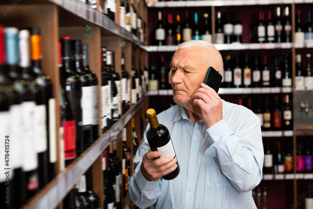 Obraz premium Senior man chooses wine and consults on a cell phone