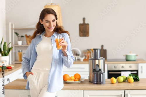 Young woman with glass of fresh fruit juice near modern juicer in kitchen