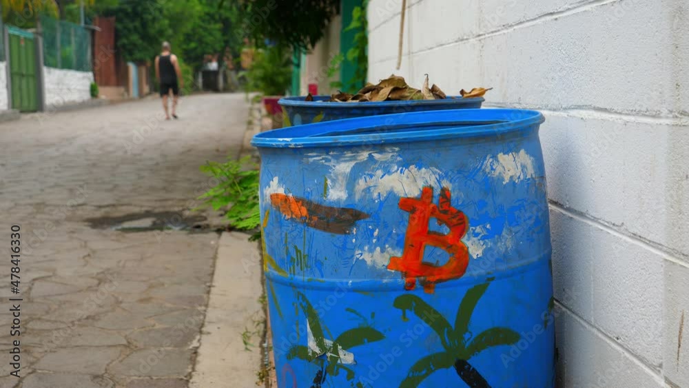 Full shot, a blue drum with a red logo of bitcoin on the bitcoin beach in El Salvador, Mexico, a person walking in the background.