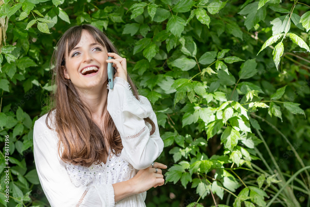 Young woman in a white dress talking amusingly on a cell phone in a park surrounded by greenery Young woman in a white dress talking amusingly on a cell phone in a park surrounded by greenery
