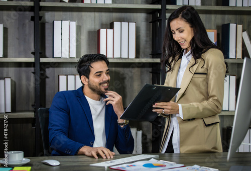 Millennial Indian Asian male successful executive businessman manager in formal suit holding clipboard discussing with female secretary businesswoman colleague at working desk in company office