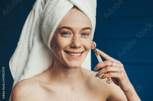 Caucasian freckled woman wrapped in towels massaging face with jade roller. Close up portrait. Smiling face. Skin care. Beauty face. photo