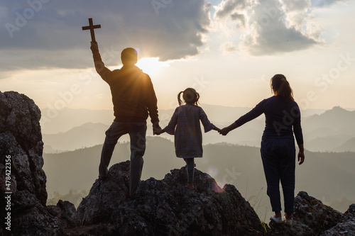 Silhouette family praying and holding Christian cross for worshipping God on mountain at sunrise background. Christian, Christianity, Religion copy space background. Easter Sunday concept: