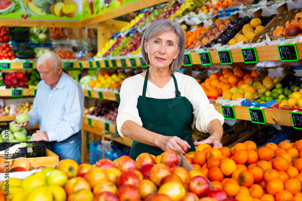 Mature woman greengrocer worker setting out goods on shelves while standing in salesroom. Old man making purchases in background.