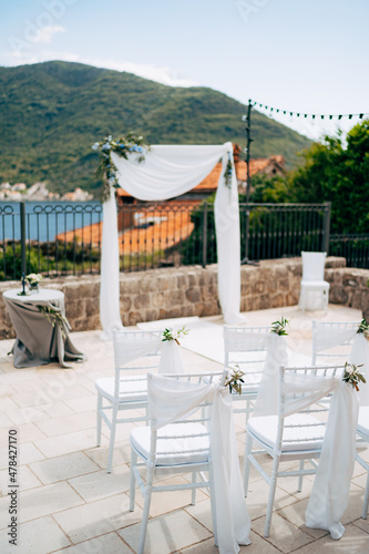 White chairs decorated with bouquets and cloth stand in front of a wedding arch in a courtyard overlooking the sea