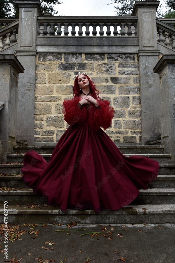  portrait of pretty  female model with red hair wearing glamorous renaissance red ballgown.  Posing in a fairytale castle location with staircases 