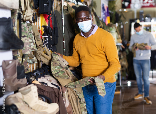 African-american man in face mask selecting outwear in military goods store.