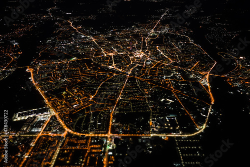 Aerial view from airplane window of buildings and bright illuminated streets in city residential area at night. Dark urban landscape at high altitude