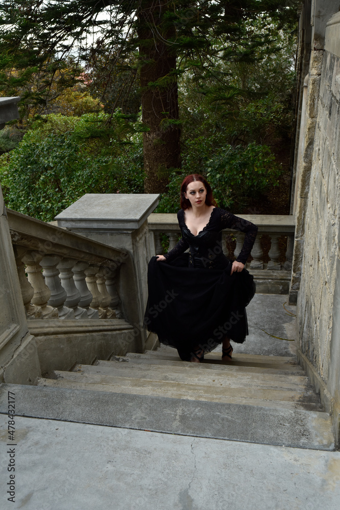  portrait of pretty  female model with red hair wearing glamorous gothic black lace ballgown.  Posing in a fairytale castle location with staircases 