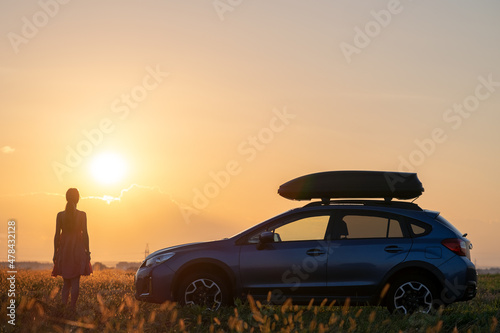 Silhouette of female driver standing near her car on grassy field enjoying view of bright sunset. Young woman relaxing during road trip beside SUV vehicle