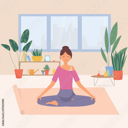 A young woman with her eyes closed is sitting in a cozy room with her legs crossed on the floor and meditating. Relaxation at home, spiritual practice, yoga and breathing exercises. 