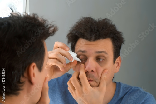 Man is dripping drops in his eyes in front of the mirror at home. He is looking at his reflection in the mirror. Eye fatigue, manager syndrome, exhaustion, stress, overwork, sleep disorder concepts. photo