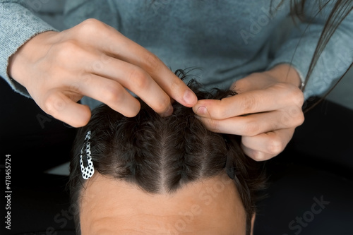 Woman is doing french braid on short men's hair, closeup view. She is doing a man's hair while sitting at home on the couch. Evening of a young married couple together.