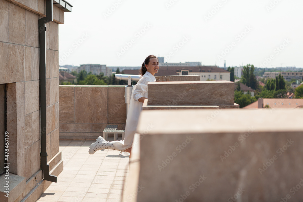 Caucasian female tourist standing on building terrace enjoying seeing landscape of metropolitan city during summer vacantion. Panoramic view of urban buildings from observation point