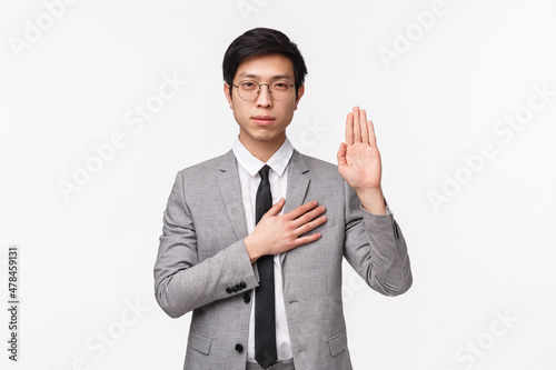 Waist-up portrait of devoted good-looking asian young man in grey suit, making promise, pledge or give oath, raise one hand and put palm on hear as being honest and sincere, white background photo