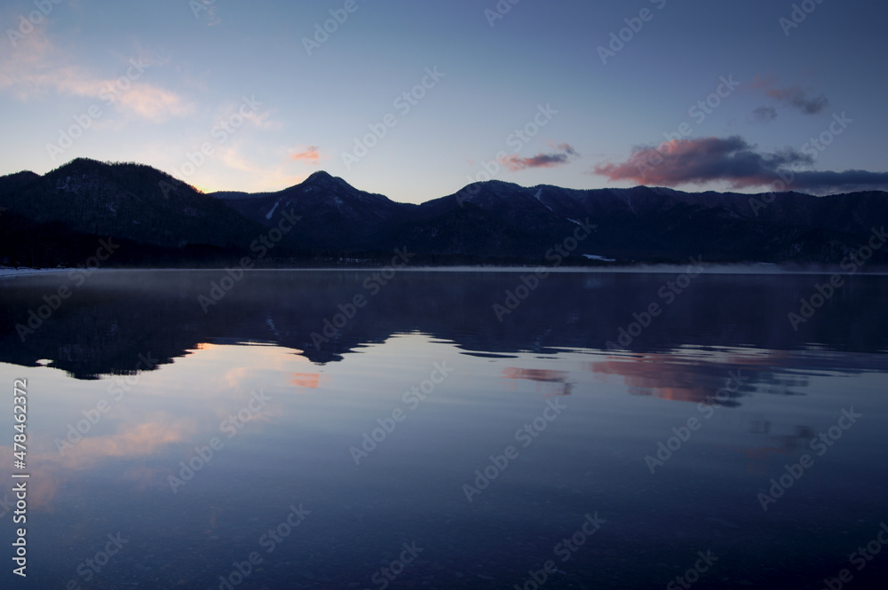 静水の湖面に山のシルエットと空を反射する黄昏の湖。日本の北海道の屈斜路湖。