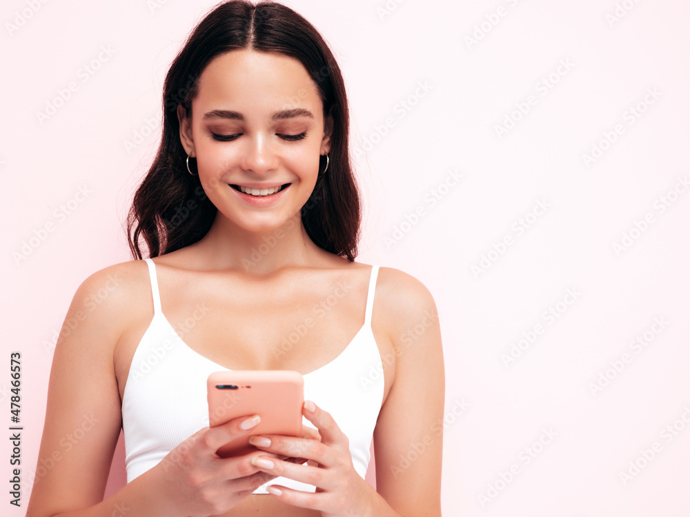 Young beautiful smiling female in trendy summer clothes. Sexy carefree woman posing near pink wall in studio. Positive brunette model looking at cellphone screen. Holding smartphone and using apps Young beautiful smiling female in trendy summer clothes. Sexy carefree woman posing near pink wall in studio. Positive brunette model looking at cellphone screen. Holding smartphone and using apps