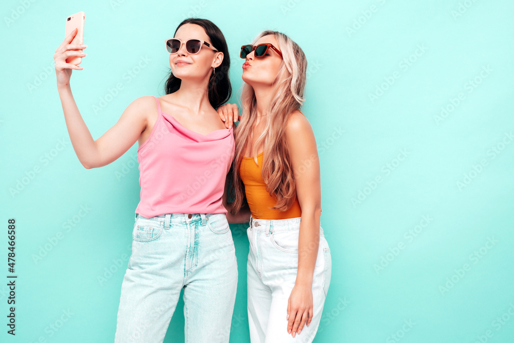 Portrait of two young beautiful smiling brunette hipster female in trendy summer clothes. Sexy carefree women posing near blue wall. Positive models having fun. Cheerful and happy. Taking selfie