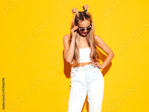 Portrait of young beautiful smiling blond female in trendy summer clothes. Sexy carefree woman posing near yellow wall in studio. Positive model having fun indoors. Cheerful and happy in sunglasses © halayalex