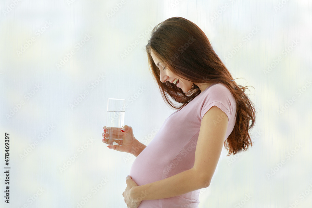 Caucasian young healthy female big belly tummy prenatal pregnant mother in casual shirt standing in front of curtain background smiling holding glass of drinking water at home in morning alone at home Caucasian young healthy female big belly tummy prenatal pregnant mother in casual shirt standing in front of curtain background smiling holding glass of drinking water at home in morning alone at home