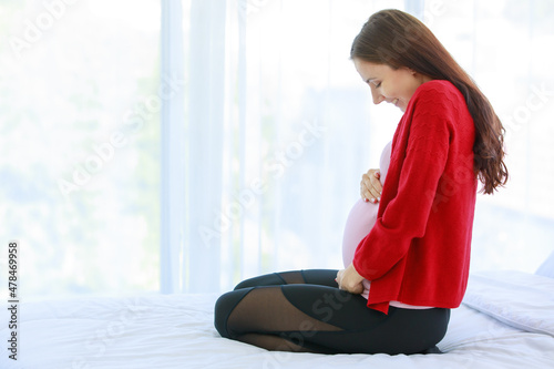 Caucasian millennial young happy female prenatal pregnant mother model in casual pregnancy outfit jacket sitting alone on bed in bedroom smiling holding hands touching on big belly tummy with love © Bangkok Click Studio