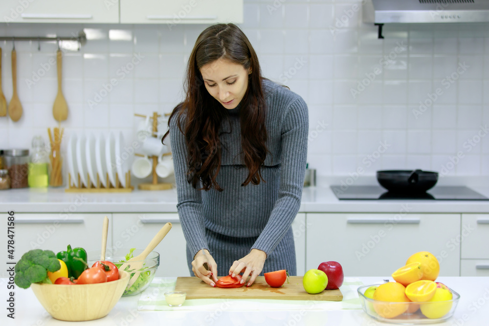 Caucasian millennial young happy sexy female prenatal pregnant mother in casual gray pregnancy dress standing smiling using knife cutting red sweet pepper on cutting board cooking vegetables salad
