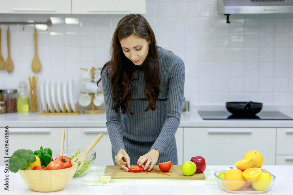 Caucasian millennial young happy sexy female prenatal pregnant mother in casual gray pregnancy dress standing smiling using knife cutting red sweet pepper on cutting board cooking vegetables salad Caucasian millennial young happy sexy female prenatal pregnant mother in casual gray pregnancy dress standing smiling using knife cutting red sweet pepper on cutting board cooking vegetables salad