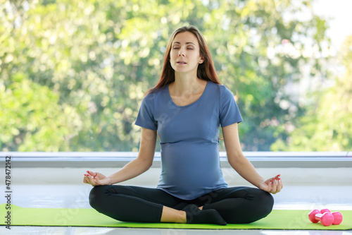 Caucasian millennial young happy female prenatal pregnant mother in casual outfit sitting crossed legs on yoga mat smiling closed eyes practicing meditation and exercise with dumbbell in living room